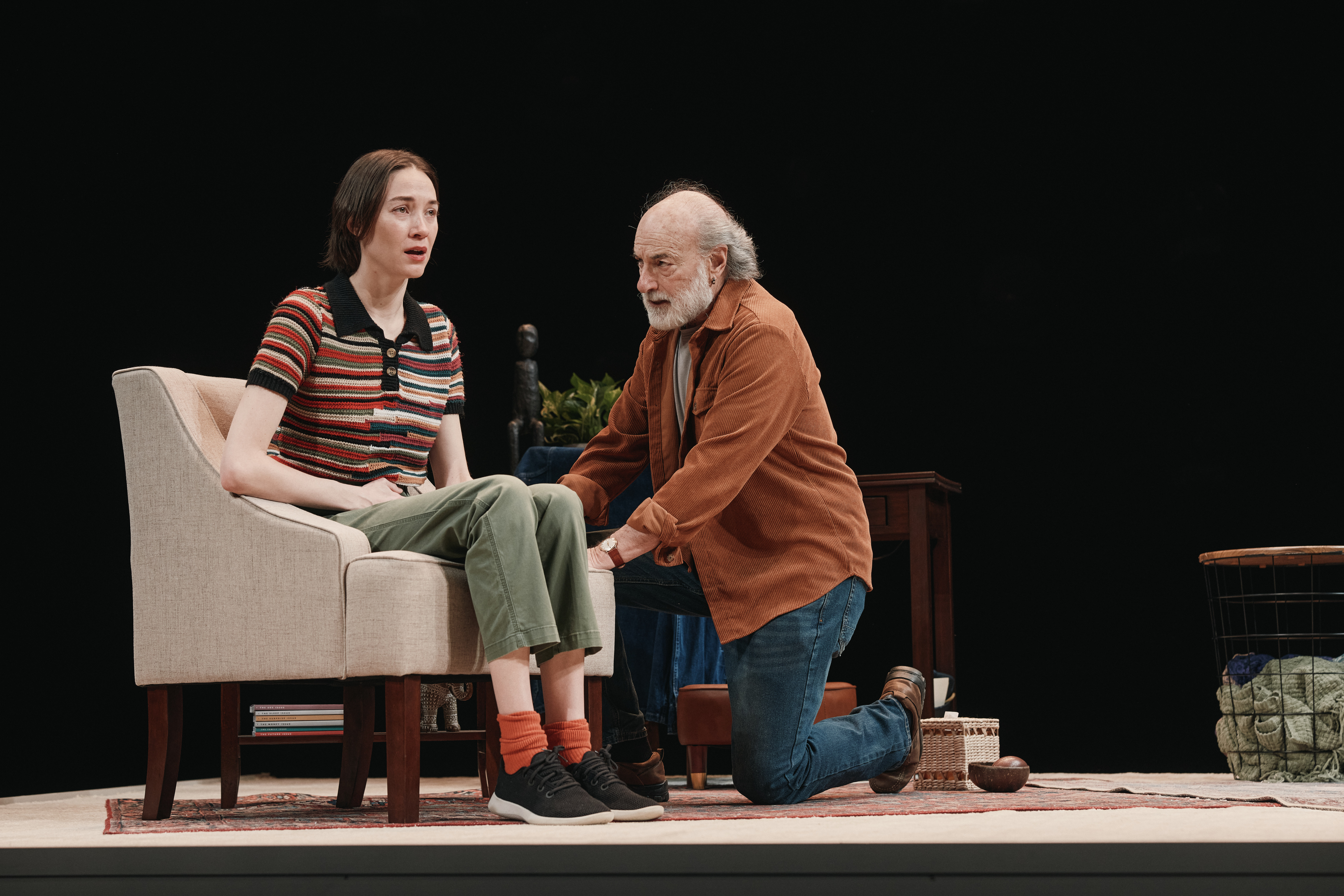 Two actors on stage with one in a chair, the other kneeling beside her